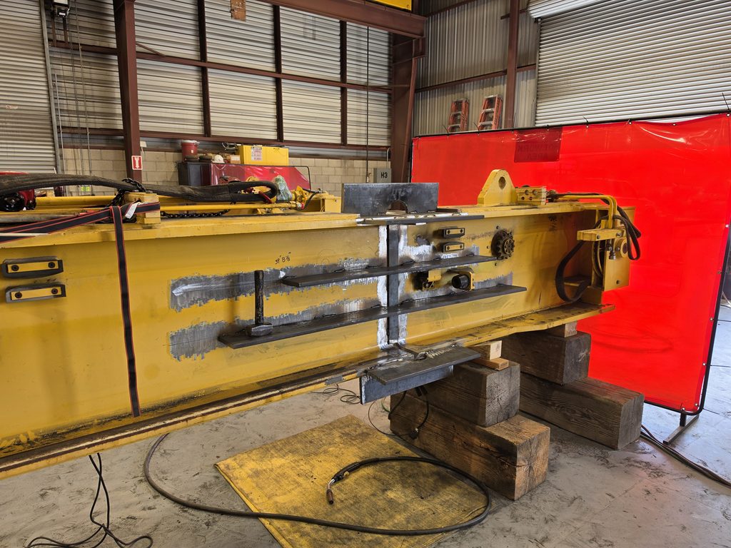 Yellow overhead crane beam on wood cribbing in the shop, with new welded steel reinforcement plates along its side