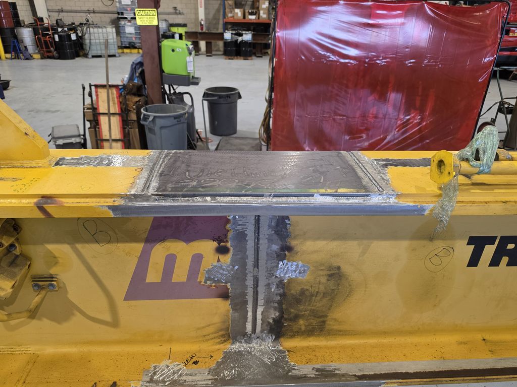 Close-up of a yellow crane boom in the Rey Welding shop with a new steel reinforcement plate and fresh structural welds across the repaired section