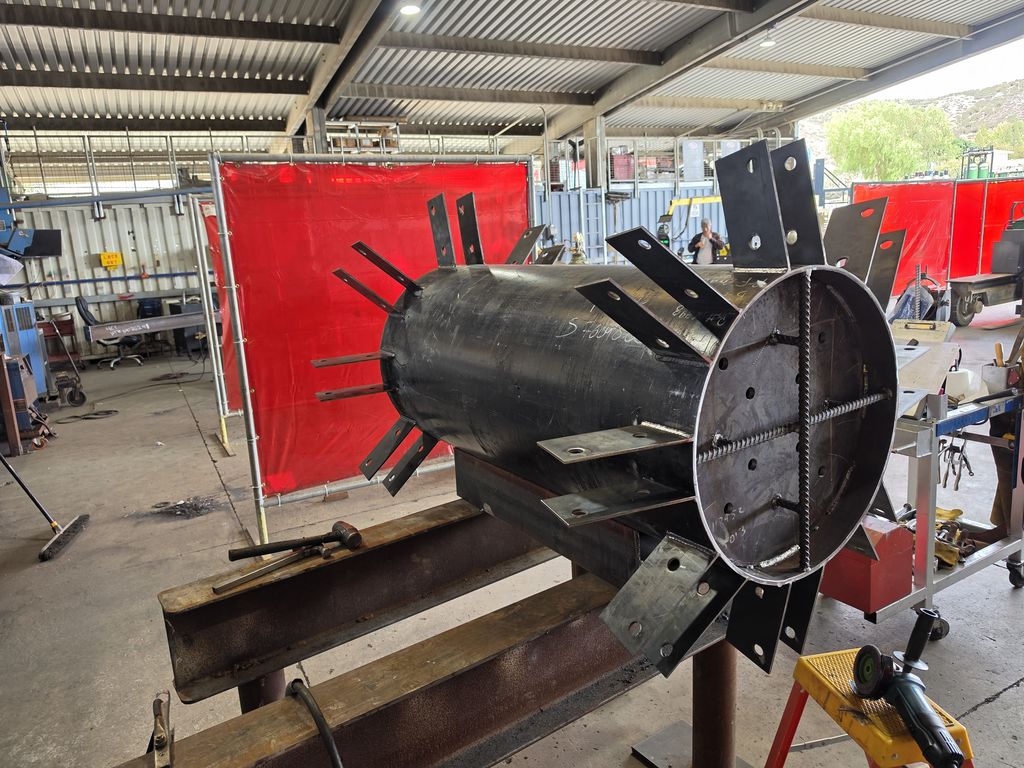 Large fabricated steel cylinder with radial welded gusset plates and a bolted end flange, on stands in the fab shop